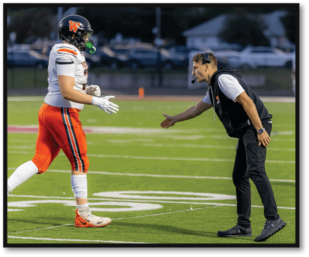 A person in orange sports pants and cleats is approaching another person in black pants and a vest who is extending a hand for a handshake on a football field. Description generated by AI
