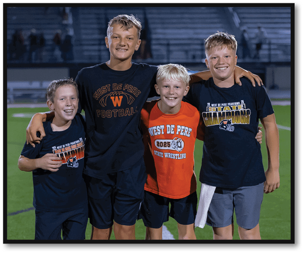 Three children smiling and standing close together on a sports field at night wearing West De Pere sports-themed shirts. Description generated by AI