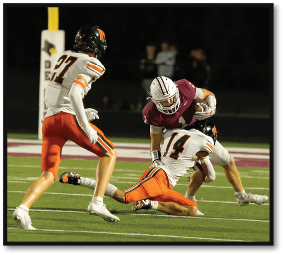 A person in a maroon sports uniform is holding a football while being tackled by a person in a white and orange uniform on a football field with another person in white and orange nearby. Description generated by AI