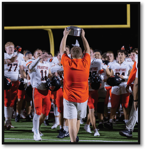 A person in an orange shirt and light shorts holds a trophy above their head while a group of people in white and red football uniforms cheer on a sports field at night. Description generated by AI