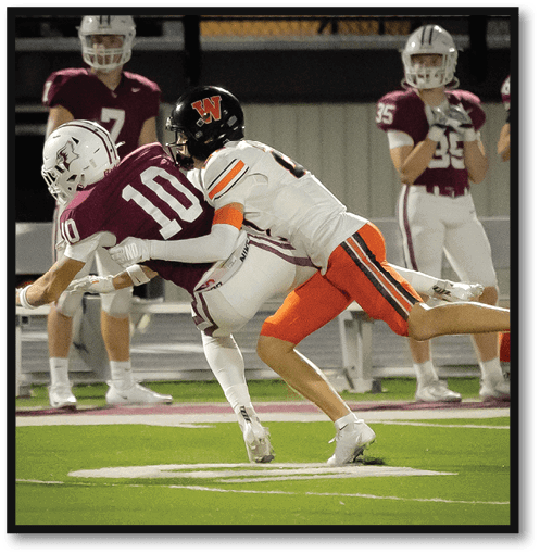 Two people in football uniforms are engaged in a tackle on a field with other players and a bench in the background. Description generated by AI