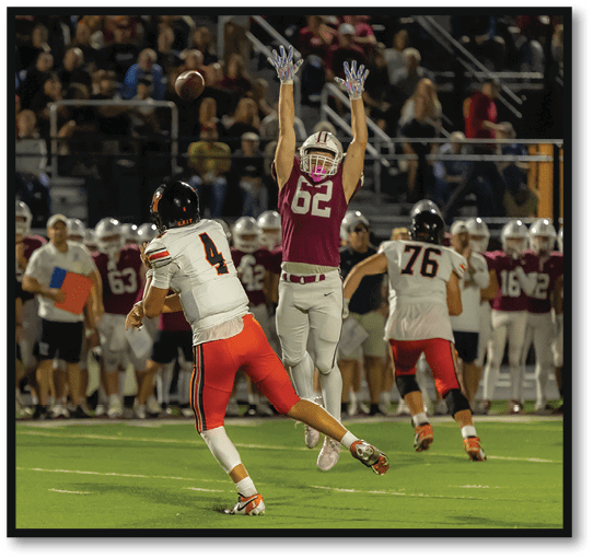 A person in a maroon football uniform jumps with arms raised attempting to block a pass from a person in a white and orange football uniform on a field with spectators in the background. Description generated by AI