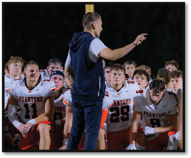 A group of people wearing football uniforms with “Phantoms" on the jerseys are kneeling and listening attentively to a person standing and gesturing in front of them on a sports field at night. Description generated by AI
