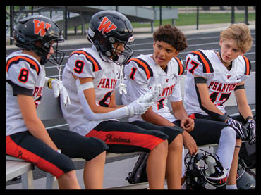 Four children in football uniforms sit on a bench at a sports field, with two wearing helmets and one holding a football helmet. Description generated by AI