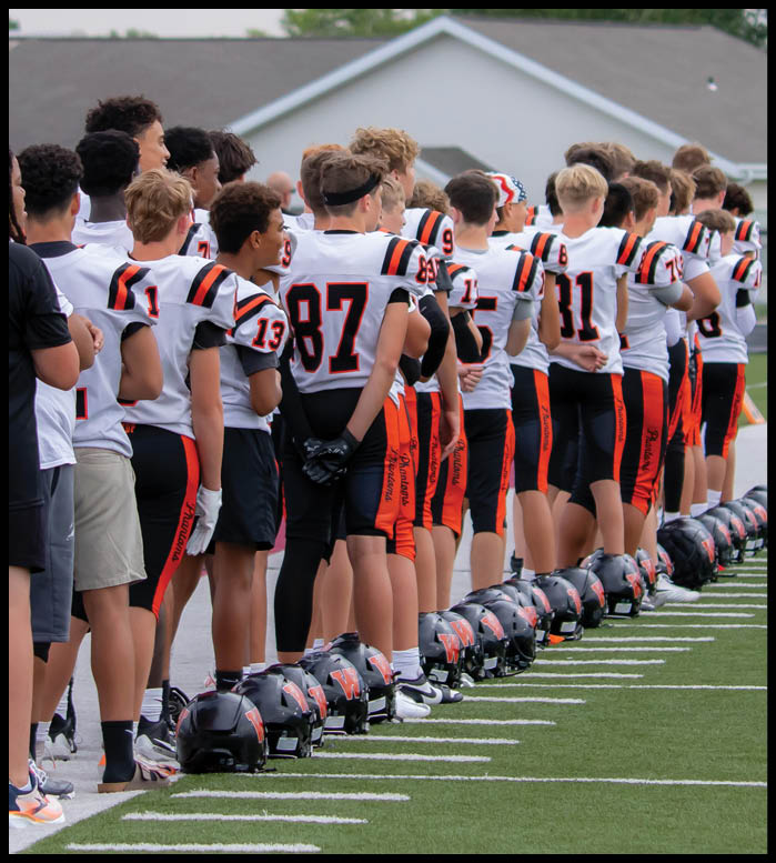 A group of people wearing football uniforms and standing in a line on a field with their helmets placed on the ground in front of them. Description generated by AI