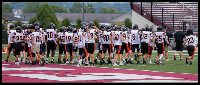 A group of people wearing football uniforms and helmets are walking together on a sports field near bleachers labeled for adults. Description generated by AI