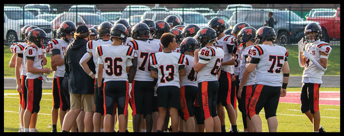 A group of people wearing football uniforms and helmets are gathered closely together on a field, appearing to be in a team huddle. Description generated by AI