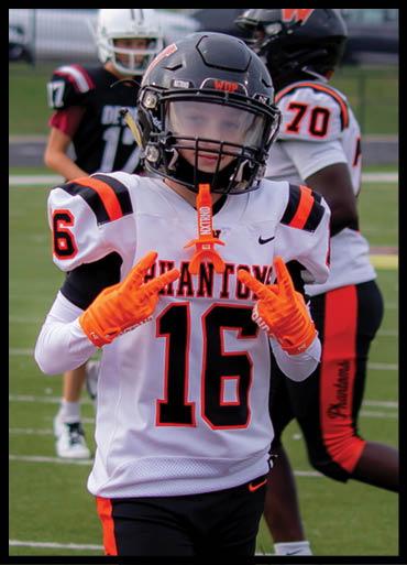 A person wearing a football uniform with the number 16 and bright orange gloves is making a hand gesture on a football field with other players in the background. Description generated by AI