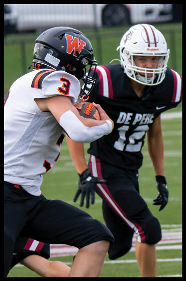 A person wearing a white and orange football uniform with the number 3 on the shoulder is holding a football while running on a grassy field. Description generated by AI