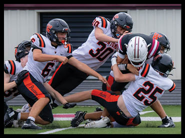 Several people wearing football uniforms are actively engaged in a play on a field, with one person in a white helmet being tackled by others in black helmets and white jerseys. Description generated by AI