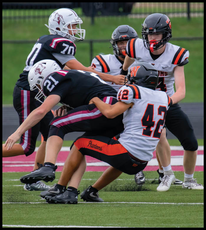 Two people in black football uniforms are being tackled by two people in white and orange football uniforms on a green field. Description generated by AI