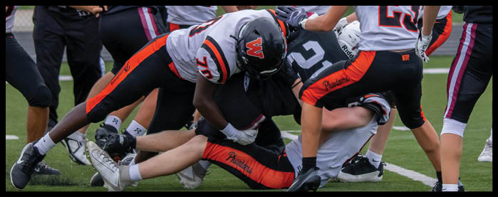 People wearing football uniforms are engaged in a play on a field with some players tackling others while referees observe nearby. Description generated by AI