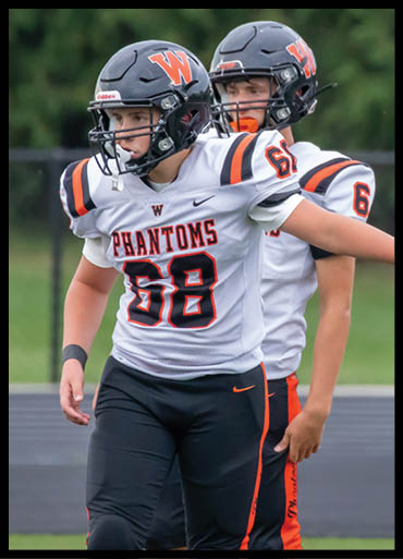 Two people wearing football helmets and white jerseys with “PHANTOMS" and the number 68 are standing on a field. Description generated by AI