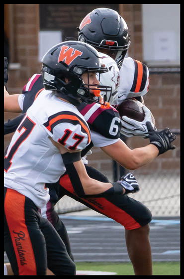 Two people in football uniforms and helmets are engaged in a play on a field, with one holding a football and the other reaching out. Description generated by AI