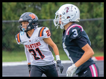 Two people wearing football helmets and uniforms are on a field, with one person in a white jersey labeled “Phantoms" and the other in a dark jersey. Description generated by AI