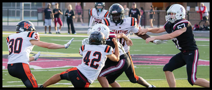 Several people wearing football helmets and uniforms are actively engaged in a play on a football field with spectators in the background. Description generated by AI