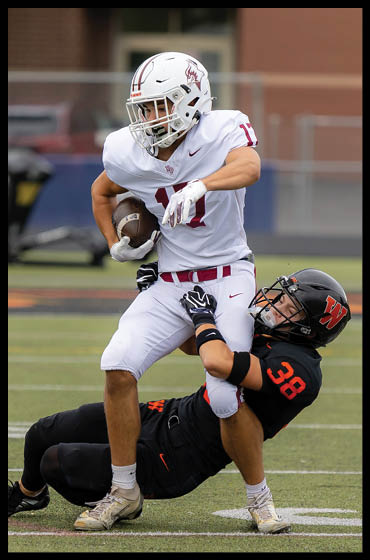 A person in a white football uniform is running with the ball while another person in a black football uniform is attempting to tackle them on a football field. Description generated by AI