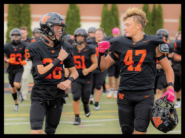Two people in black and orange football uniforms are running on a field with others in similar uniforms in the background. Description generated by AI