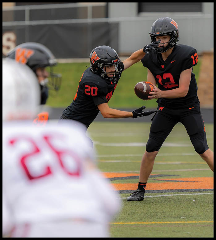 Two people in black football uniforms are engaged in a play on a field while a person in a white uniform with the number 26 is in the foreground. Description generated by AI