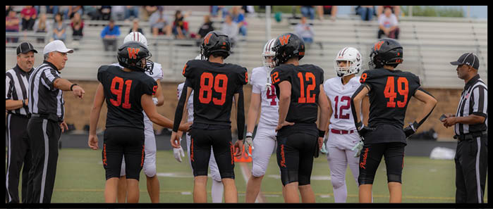 Five people in black football uniforms with orange numbers stand on a field facing three people in white football uniforms while two referees in black and white striped shirts stand nearby. Description generated by AI