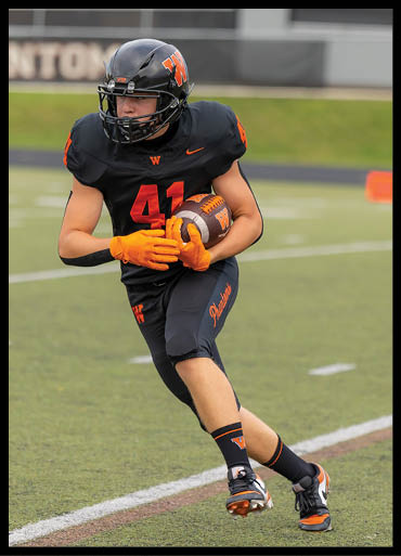 A person wearing a black football uniform with orange accents is running on a field while holding a football. Description generated by AI