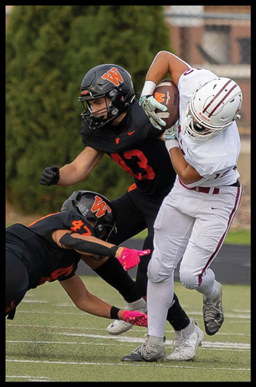 A person in white sports gear is running with a football while two people in black uniforms attempt to tackle them on a football field. Description generated by AI