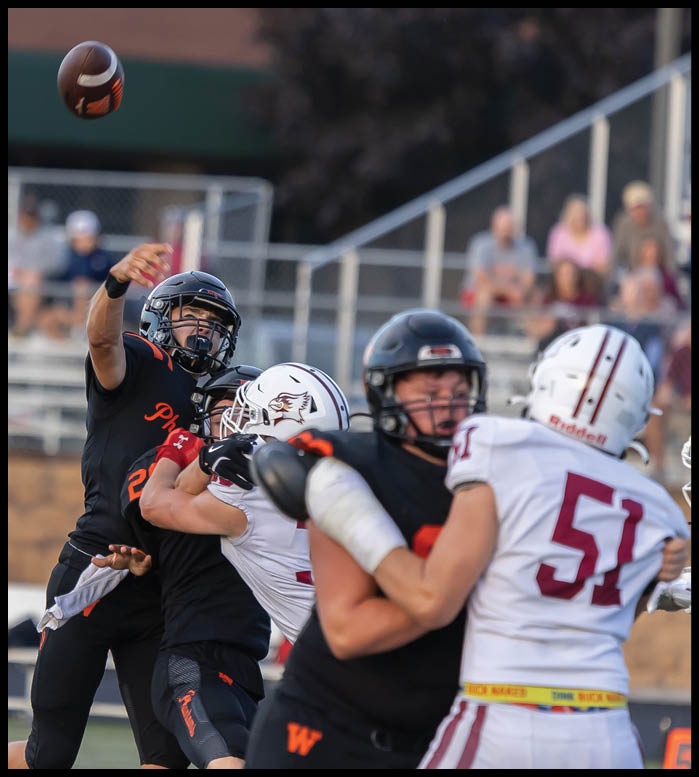 A person in black sports gear is throwing a football while being closely defended by another person in white sports gear during a game on a field with blurred spectators in the background. Description generated by AI