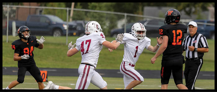 People in football uniforms are actively reaching for a football in midair on a grassy field with a referee observing nearby. Description generated by AI