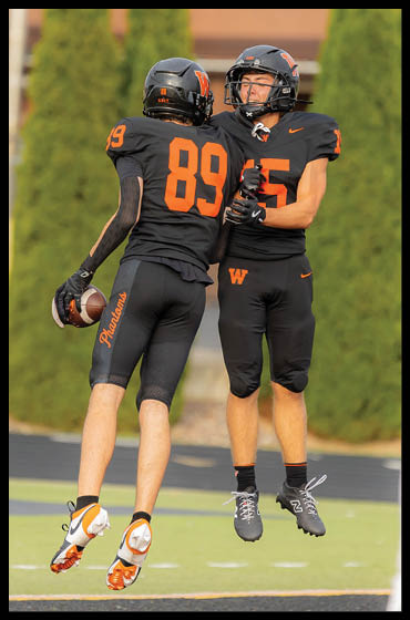 Two people in black football uniforms with orange numbers jump and celebrate on a field while a person in a white uniform with red numbers stands nearby. Description generated by AI