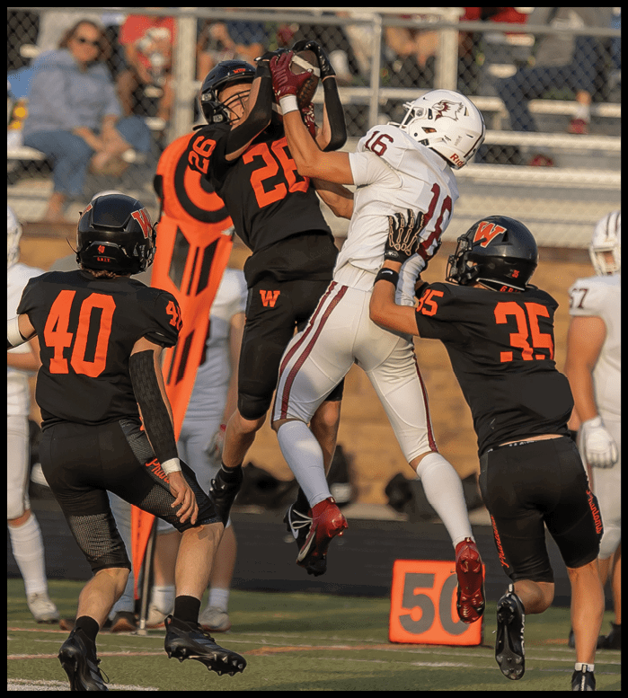Four people in football uniforms are jumping and reaching for a football in midair on a field with a crowd in the background. Description generated by AI