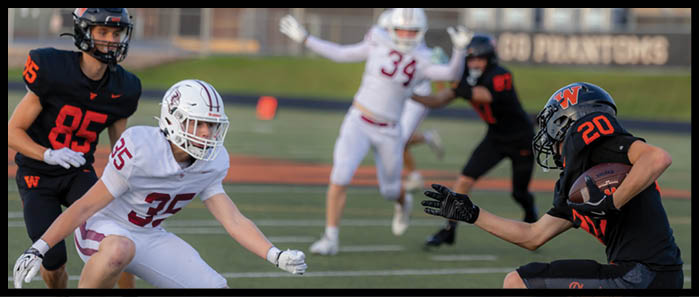 Four people in football uniforms are actively engaged in a play on a field, with one person holding the ball and others positioned to block or tackle. Description generated by AI