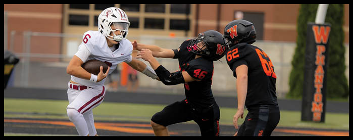 A person in a white sports uniform is running with a football while two people in black uniforms attempt to block or tackle them on a football field. Description generated by AI