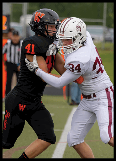 Two people in football uniforms are engaged in a physical play on a field, with one wearing black and the other wearing white. Description generated by AI