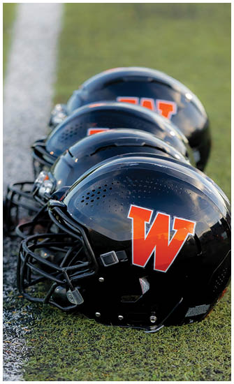 A close-up view of stacked black football helmets with orange lettering on a blurred green background. Description generated by AI