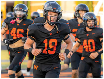 A person wearing a black football uniform with the number 19 in orange is running on a field with other similarly dressed players in the background. Description generated by AI