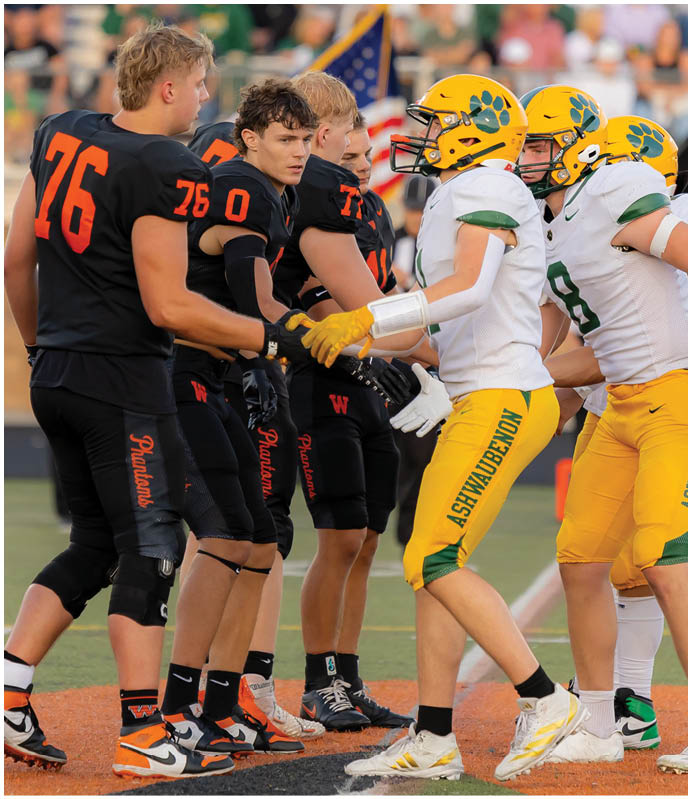People in football uniforms from two teams are shaking hands on a field, with one team wearing black and orange and the other wearing white and yellow. Description generated by AI