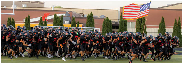 A large group of people wearing black sports uniforms and helmets are gathered closely together on a field with an American flag waving in the background. Description generated by AI