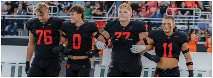 Four adults in black and red football uniforms are walking arm in arm on a football field. Description generated by AI