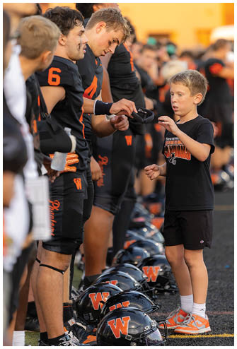 Adults wearing black sports uniforms with orange details stand closely together, with one person focused on adjusting a black wristband. Description generated by AI
