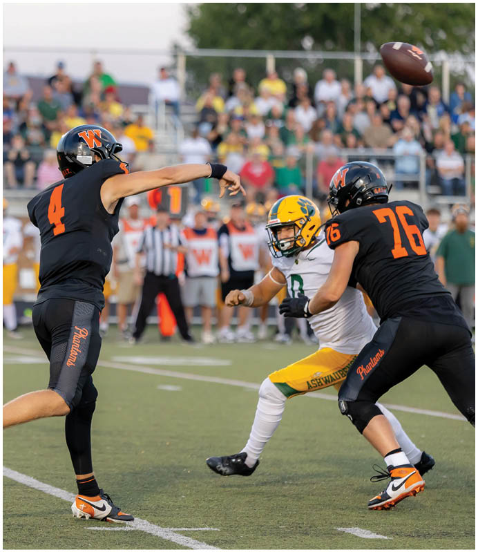 A person wearing a black sports uniform with the number 4 is throwing a football while another person in a white and yellow uniform prepares to block on a football field with a crowd in the background. Description generated by AI