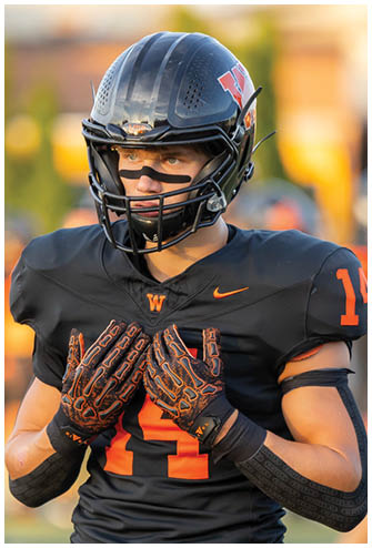 A person wearing a black football helmet, black jersey with an orange “W" and Nike logo, and black and orange gloves is looking intently to the side. Description generated by AI