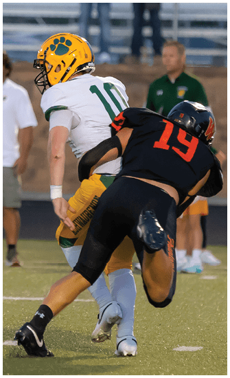 A person in a yellow and green football uniform is crouched on a field near a marker showing the number 30 while another person in casual clothing stands blurred in the background. Description generated by AI