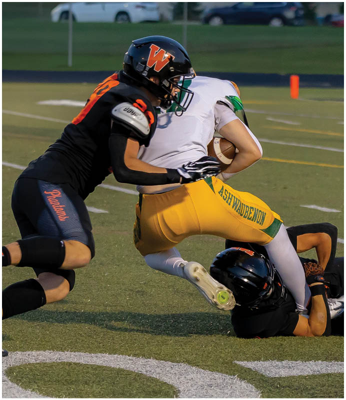 A person in yellow and green sportswear is being tackled by two people in black uniforms during a football game on a field. Description generated by AI
