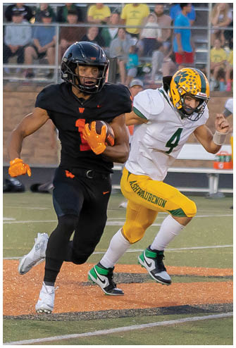 Two people in yellow and white football uniforms are actively playing on a field with spectators and officials in the background. Description generated by AI