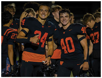 Two adults wearing black sports jerseys with orange numbers smile and pose together on a field at night. Description generated by AI