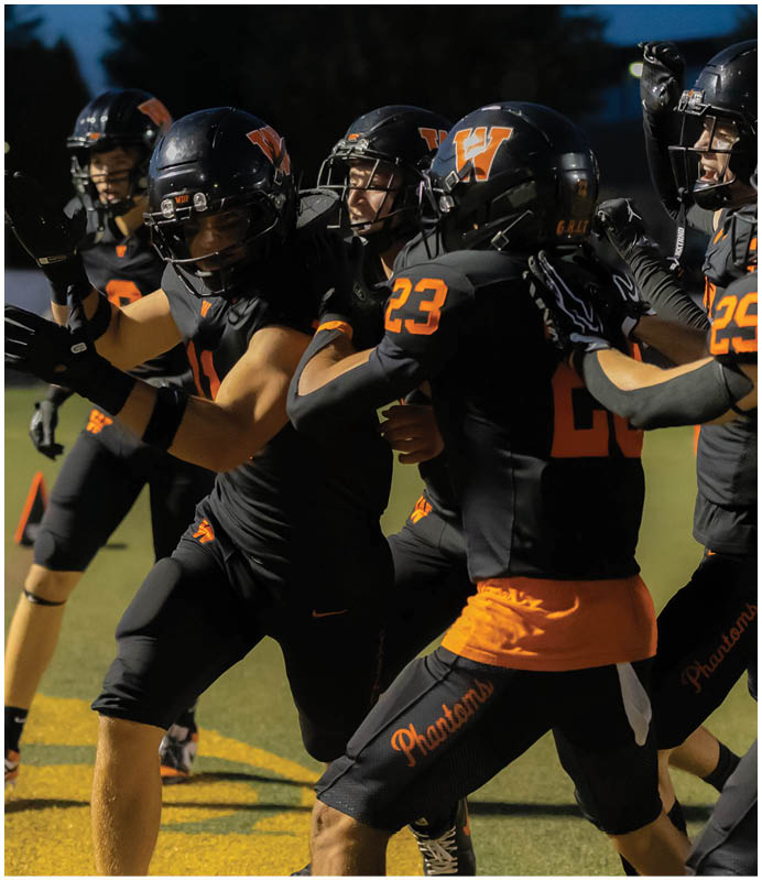A group of people wearing black and orange football uniforms with helmets are celebrating together on a field. Description generated by AI