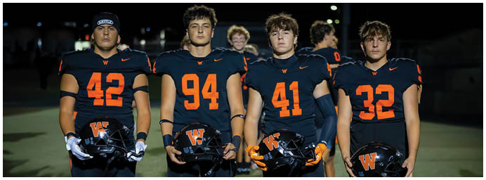 Four people wearing dark football uniforms with orange numbers stand side by side holding helmets in a nighttime outdoor setting. Description generated by AI