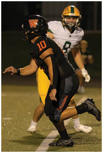 An adult wearing a black football uniform with the number 10 and the word Phantom on the pants is in a crouched position on a football field. Description generated by AI