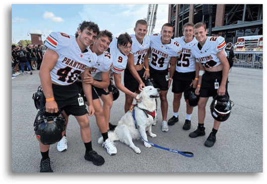 A group of people wearing matching sports jerseys are smiling and gathered around a white dog wearing a harness outdoors. Description generated by AI