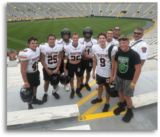 A group of people wearing football jerseys and helmets stand together smiling in a large empty stadium with green field and tiered seating. Description generated by AI